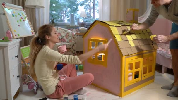 junge Familie mit Kind baut und bemalt gemeinsam Spielzeugkartonhaus.