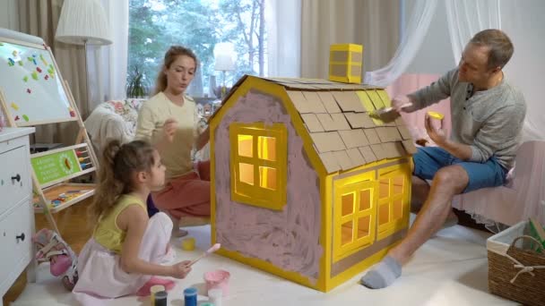 Junge Familie mit Kind baut und bemalt gemeinsam Spielzeugkartonhaus. — Stockvideo