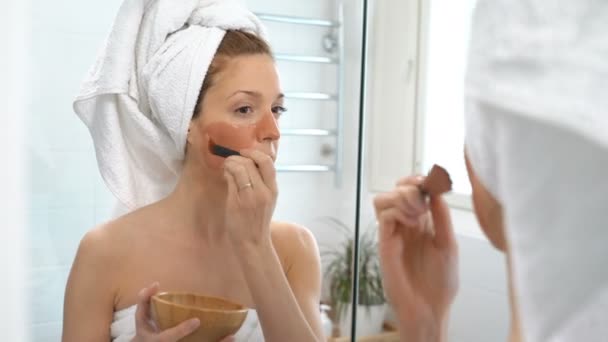 A young woman with a white towel put on her face a brown moisturizing mask — Stock Video