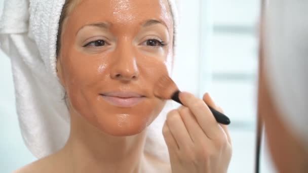 Een jonge vrouw met een witte handdoek zetten van haar gezicht een bruin hydraterende masker — Stockvideo