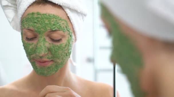 Μια νεαρή γυναίκα με μια άσπρη πετσέτα βάζουμε στο πρόσωπό της ένα πράσινο ενυδατική μάσκα — Αρχείο Βίντεο