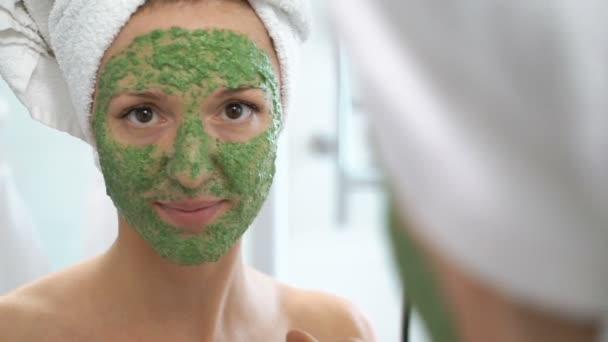 Μια νεαρή γυναίκα με μια άσπρη πετσέτα βάζουμε στο πρόσωπό της ένα πράσινο ενυδατική μάσκα — Αρχείο Βίντεο
