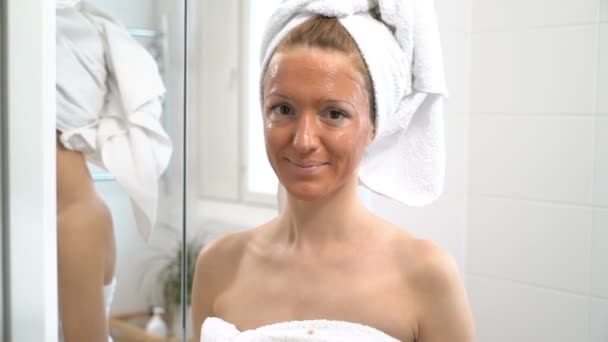 A young woman with a white towel put on her face a brown moisturizing mas — Stock Video