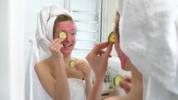 Jonge vrouw met een roze hydraterende gezichtsmasker met plakjes komkommer speelt — Stockvideo