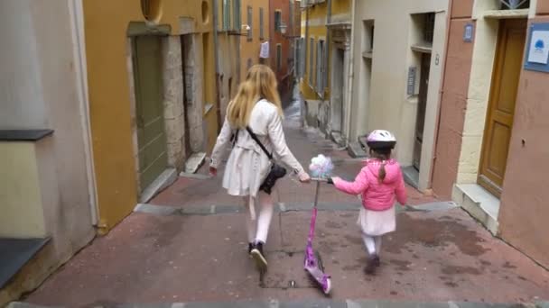 Mother and her daughter walk along the narrow street of the old European city with scooter — Stock Video