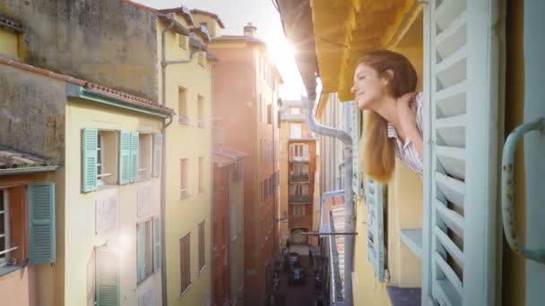 Молодая женщина смотрит в окно на средневековую улицу, улыбается и машет рукой. — стоковое видео