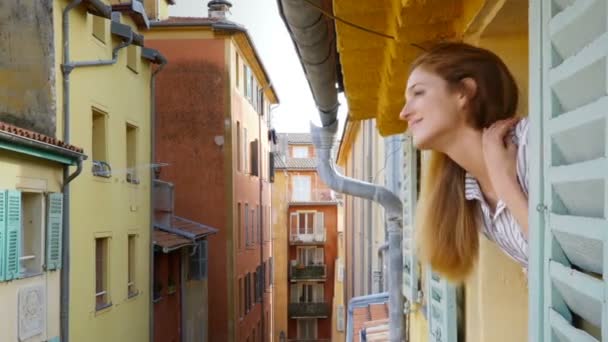 Una joven mirando por la ventana a una calle medieval sonriendo y agitando la mano — Vídeos de Stock