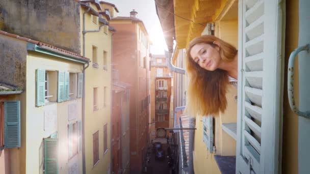 A young woman looking out of the window to a medieval street smiling and waving her hand — Stock Video