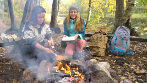 Female friends cooking traditional pancakes over an open fire in the camp outdoor during a hike — ストック動画
