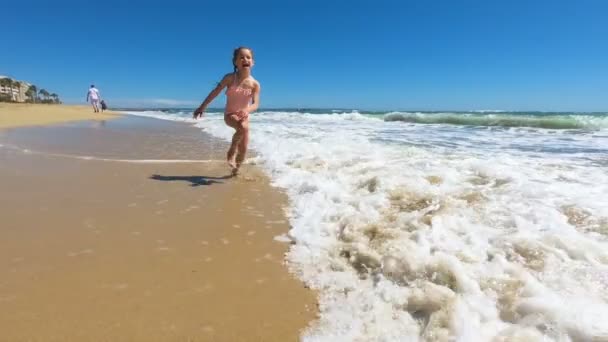 Liten flicka bär rosa baddräkt som springer på stranden barfota — Stockvideo