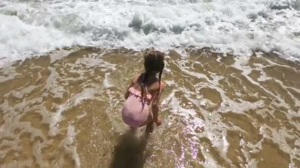 Szczęśliwa dziewczynka w różowym stroju kąpielowym skacze i bawi się falami na plaży — Wideo stockowe