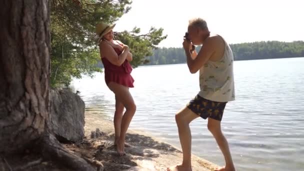 Homem idoso tirando foto de mulher sênior atraente com câmera vintage na praia . — Vídeo de Stock