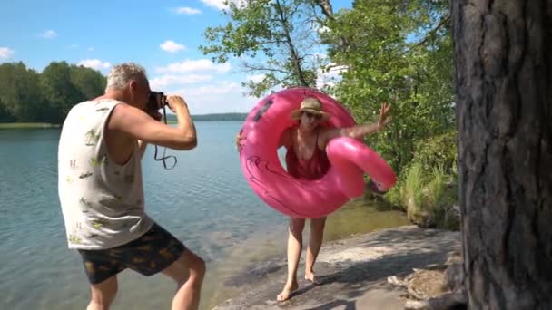 Homem idoso tirando foto de mulher sênior atraente com câmera vintage na praia . — Vídeo de Stock