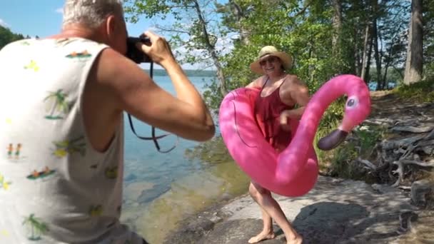 Homem idoso tirando foto de mulher sênior atraente com câmera vintage na praia . — Vídeo de Stock