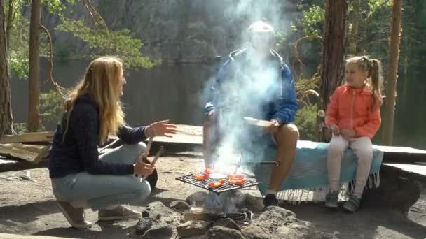 Una pareja con un niño a la parrilla en el bosque a orillas del lago — Vídeo de stock