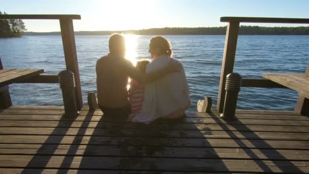 Couple with child sitting on a wooden jetty at sunset near the lake — Stockvideo