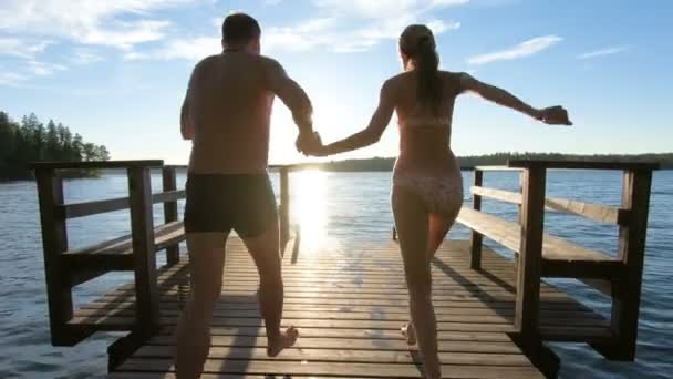 Пара бежит по деревянному пирсу и прыгает в озеро — стоковое видео