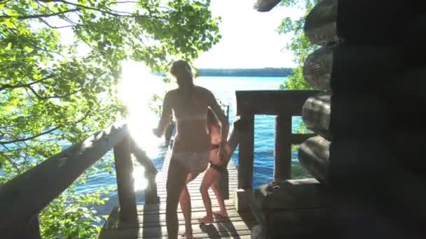 Женщина бежит вниз по лестнице от сауны к деревянному пирсу, чтобы прыгнуть в озеро — стоковое видео