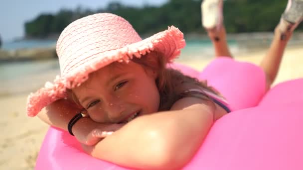 Una niña pequeña con un sombrero de paja rosa acostada en un sofá inflable en una playa — Vídeos de Stock