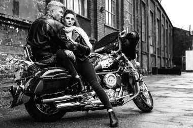 Esmer kız, gri saçlı, deri ceketli, motosikletli bir adamla oturuyor.