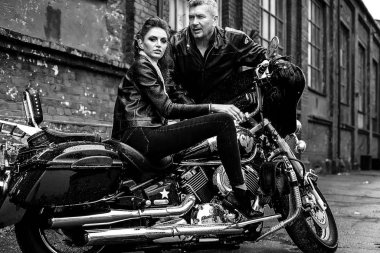 Esmer kız, gri saçlı, deri ceketli, motosikletli bir adamla oturuyor.