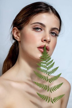 Beyaz arka planda yeşil eğreltiotu olan esmer bir kadının portresi.