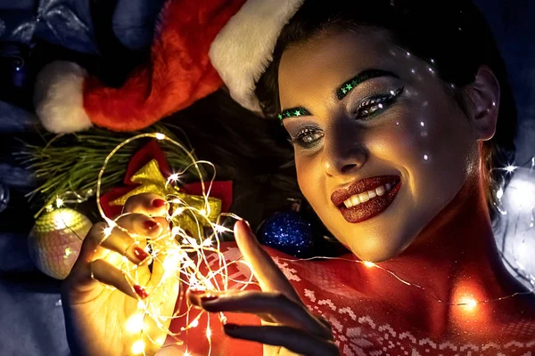 Brunetka dívka v klobouku Santa Claus, v noci na modrém pozadí, drží světla v rukou a usmívá se roztomilý. — Stock fotografie