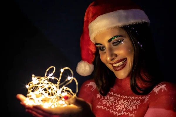 En brunett flicka i en jultomte hatt, på natten på en blå bakgrund, håller ljus i händerna och ler sött. — Stockfoto