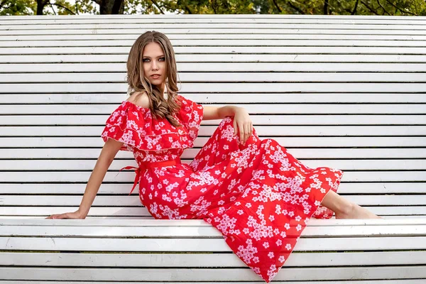 Güzel, genç bir kız, model, uzun saçlı, kırmızı elbiseli, kahverengi saçlı bir kadın bankta oturuyor ve rahat, şaşkın bir bakışla poz veriyor..