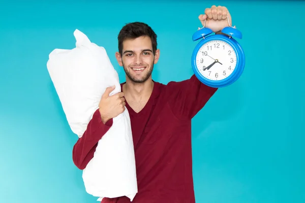 young man with pajamas and alarm clock with pillow isolated on color background