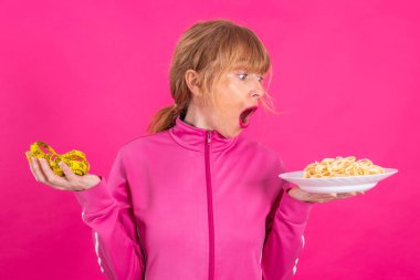 woman with stress expression looking at measuring tape and plate of pasta or hydrates with sportswear isolated on pink background