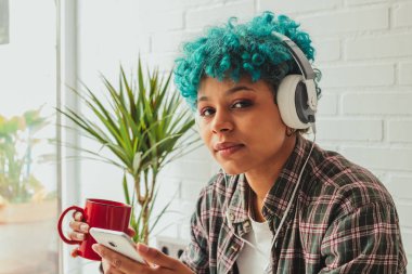 Kulaklıklı ve cep telefonlu bir kız kahve içiyor ve evde müzik dinliyor.