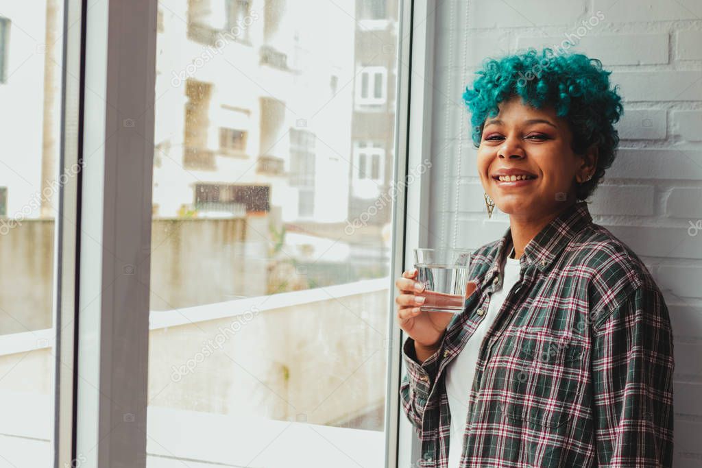 young woman or blue-haired african american girl in the apartment with glass of water at the window