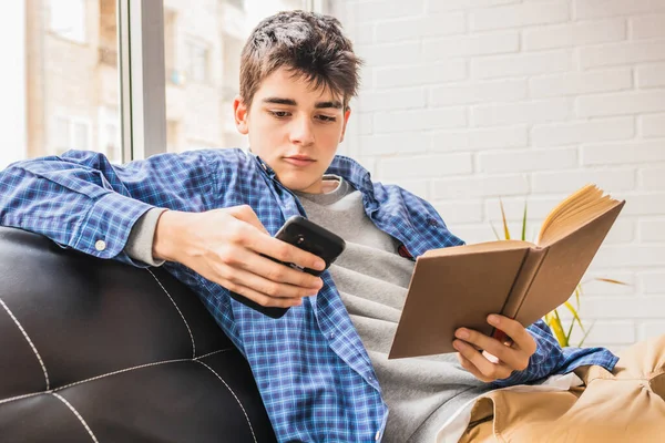 家やアパートで携帯電話や本を持つ若い10代の男の子 — ストック写真
