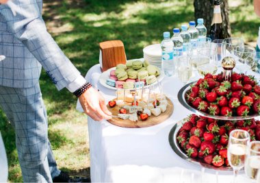 Açık mavi takım elbiseli bir adam yemek masasından çilekli, peynirli ve bardaklı yiyecek alır.