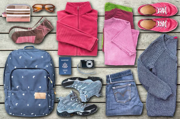 View from above of unisex travel essentials such as a backpack, shoes, clothes, a digital camera, sunglasses, a cosmetic bag, a passport (USA), organized to fill the space on the wooden floor