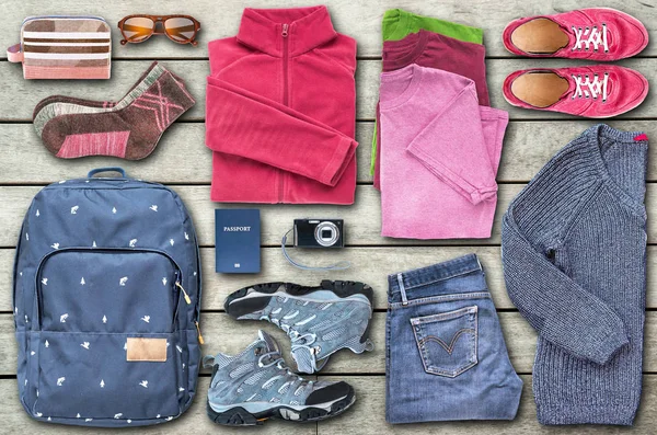 A view from above of unisex travel essentials such as a backpack, shoes, clothes, a digital camera, sunglasses, a cosmetic bag and a passport, organized to fill all the space on the wooden floor