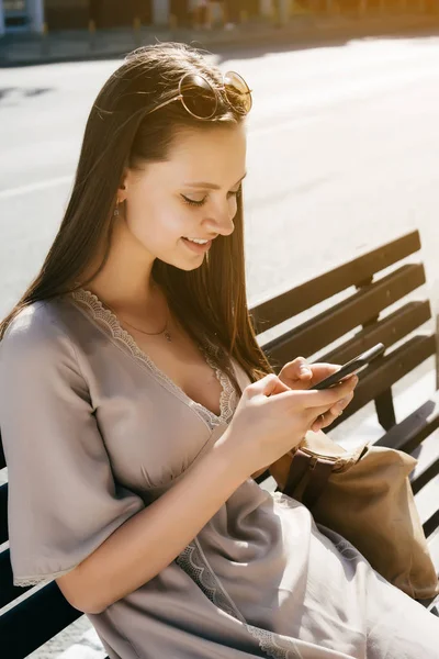 Mujer de moda sentada en el banco y el uso de teléfono inteligente, sonriendo, w — Foto de Stock