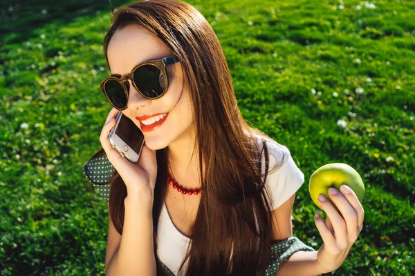 Mutlu gülümseyen kadın güneş gözlüğü, cep telefonunu kullanma ve elma tutar