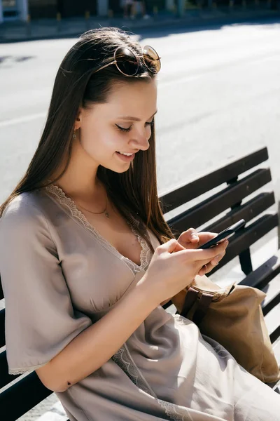 Mujer de la moda sentada en el banco y el uso de teléfono inteligente, sonriendo, viendo vídeo — Foto de Stock