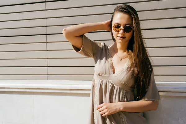 Yuvarlak gözlüklü bir kız güneşin altında sokakta duruyor.