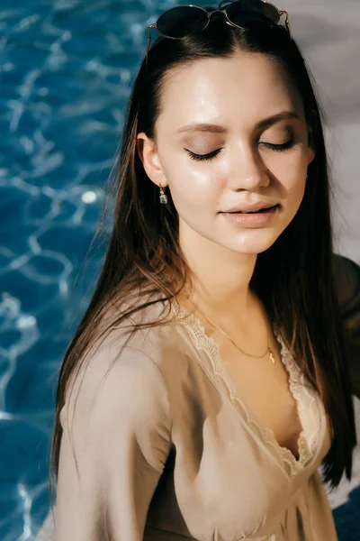 Joven mujer de moda sentada cerca de la piscina, sonriendo con los ojos cerrados — Foto de Stock