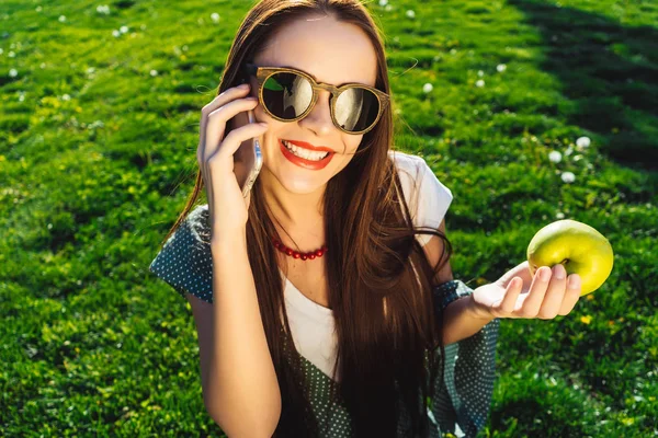 Feliz mujer sonriente en gafas de sol utilizar el teléfono celular y sostiene la manzana — Foto de Stock