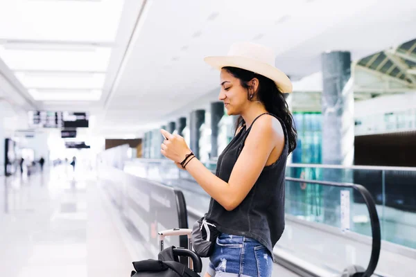 Airport woman on smart phone at gate waiting in terminal. Air travel concept with young casual hipster woman standing with carry-on hand luggage trolley. Beautiful young mixed race female — Stock Photo, Image