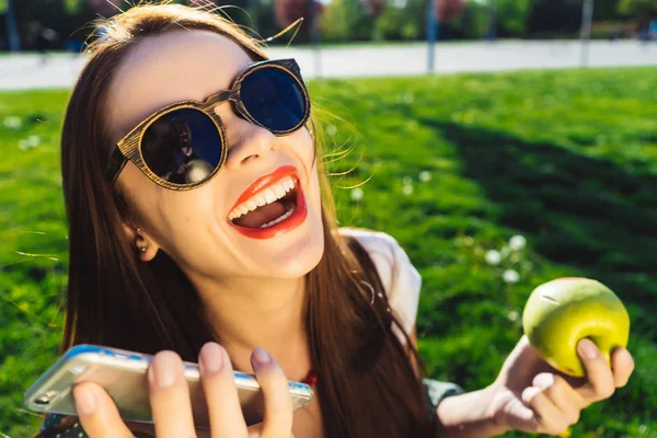 Chica brillante en gafas con lápiz labial rojo sostiene manzana y teléfono en la naturaleza — Foto de Stock