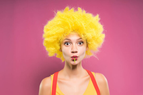 Забавная девушка в жёлтом парике смотрит в камеру и ест чупа-чуп.
