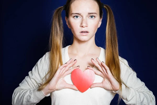 Menina séria em um fundo azul escuro detém um coração de papel — Fotografia de Stock