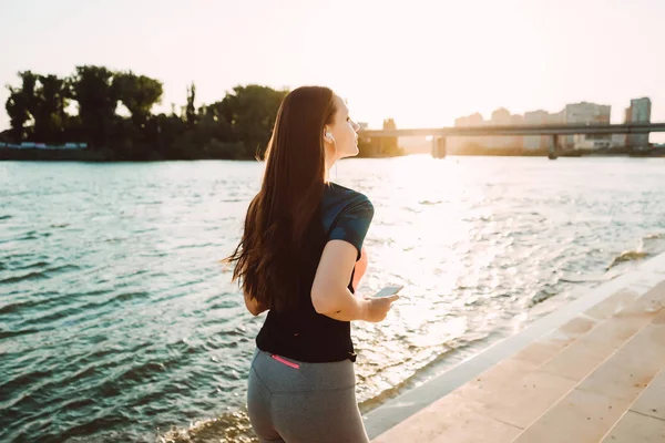 Sportig tjej i hörlurar gör en kväll kör vid solnedgången vid floden med en telefon i händer — Stockfoto