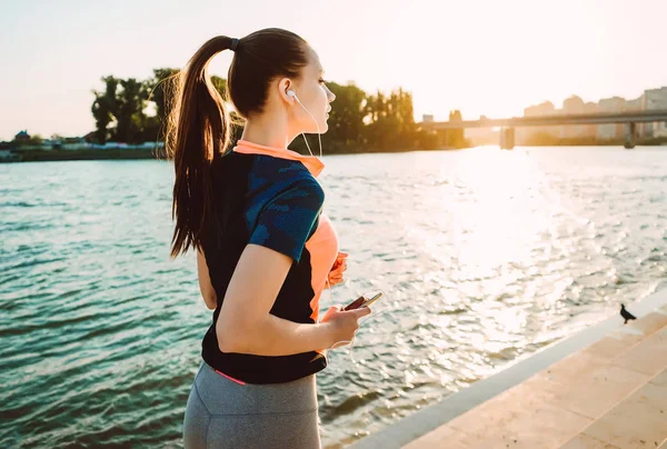 Sportig tjej i hörlurar gör en kväll kör vid solnedgången vid floden med en telefon i händer — Stockfoto