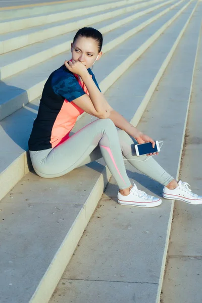 Meisje in een sport-pak zit op de trappen en luistert naar muziek — Stockfoto